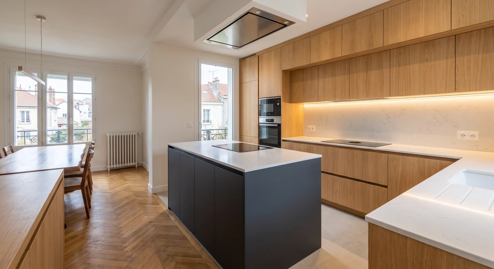 Photographie d'une cuisine moderne sur mesure complètement aménagée et finalisée, montrant l'intégration harmonieuse de tous les éléments : façades en bois naturel ou finition mate, plan de travail assorti, électroménager encastré de qualité (four, plaque induction, hotte), crédence intégrée dans la continuité, éclairage sous-meuble LED discret, poignées assorties, sol carrelé ou parqueté, avec si possible un détail de l'agencement environnant (mur adjacent, transition vers salle à manger ou salon). L'image doit montrer la cohérence d'ensemble et le finition aboutie d'un projet global maîtrisé.