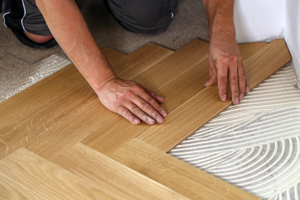 Photographie réaliste d'un professionnel artisan en tenue de travail (menuisier agenceur) dans un atelier ou sur un chantier de rénovation intérieure en cours. L'image montre le professionnel prenant des mesures ou inspectant un espace résidentiel semi-vide en rénovation (murs apparents, sol partiellement aménagé). En arrière-plan ou dans un encart incrustée, visualisation partielle d'une cuisine moderne finalisée ou d'un meuble sur mesure. L'image transmet l'expertise, le suivi de projet de la conception à la réalisation, et l'approche "interlocuteur unique" menée par Into The Wood. Lumière naturelle, cadre professionnel et épuré, sans texte ni logo.